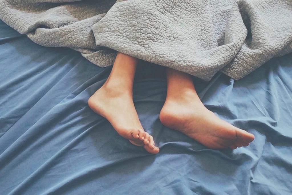 Bare feet poking out from under a grey blanket on blue sheets.